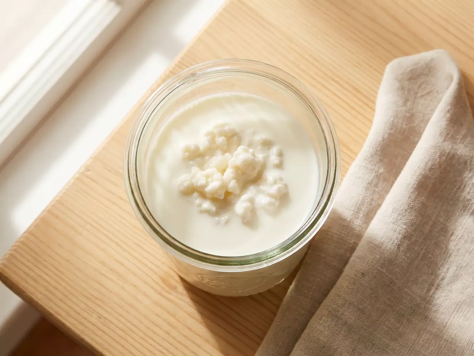 Tarro de cristal con k&eacute;fir de leche fermentando sobre madera junto a un pa&ntilde;o de lino.