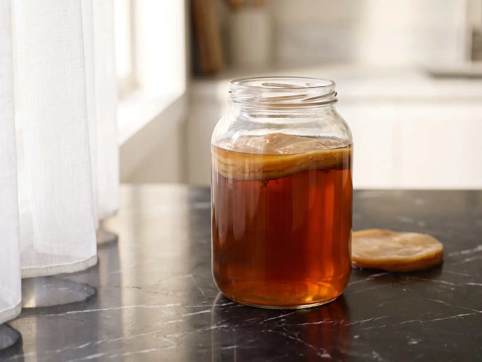 Tarro con SCOBY y t&eacute; &aacute;mbar profundo sobre m&aacute;rmol negro mate con reflejo de cortina blanca.