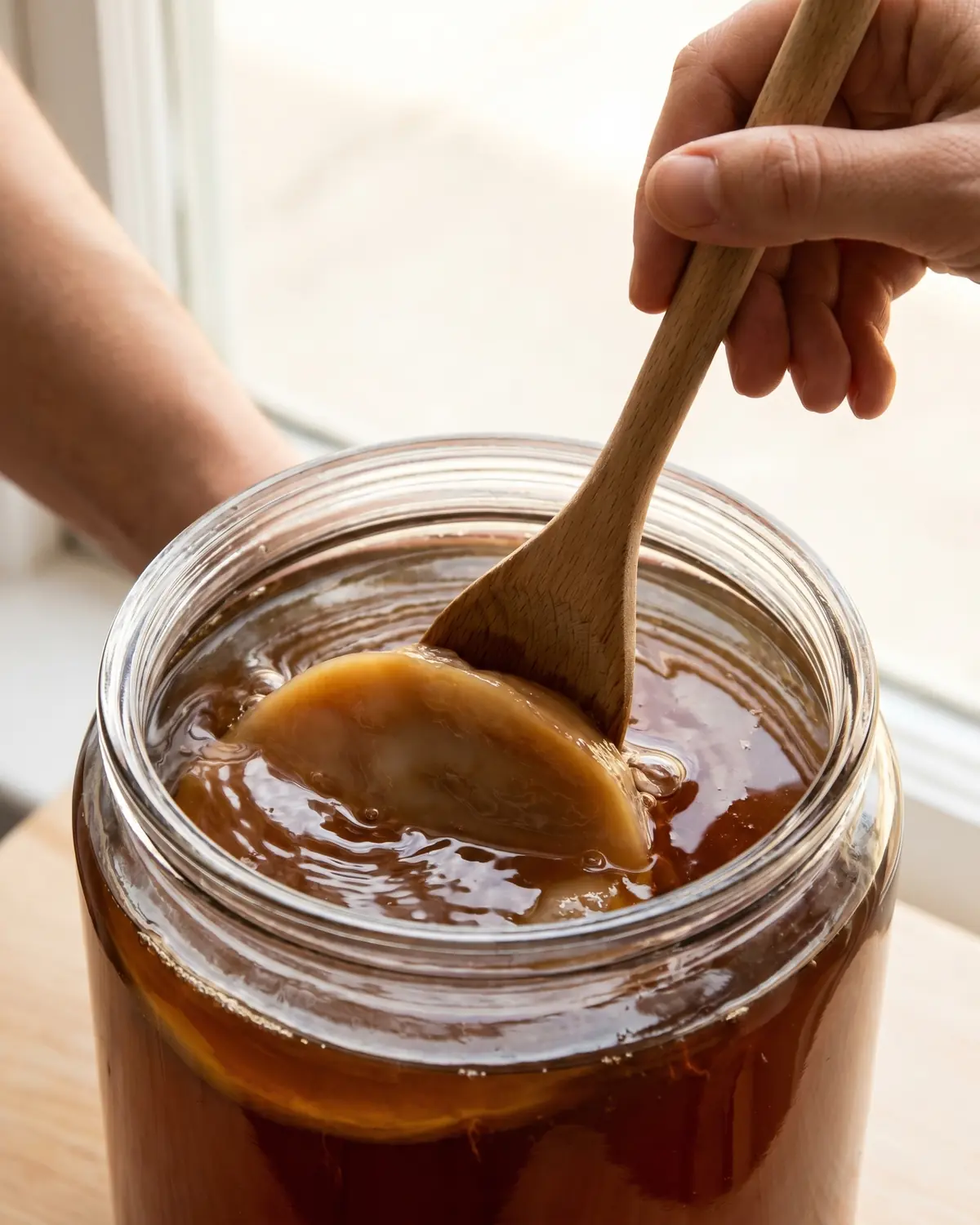 kombucha lidl Cuchara de madera empujando suavemente un SCOBY bajo té ámbar durante la preparación de kombucha casera