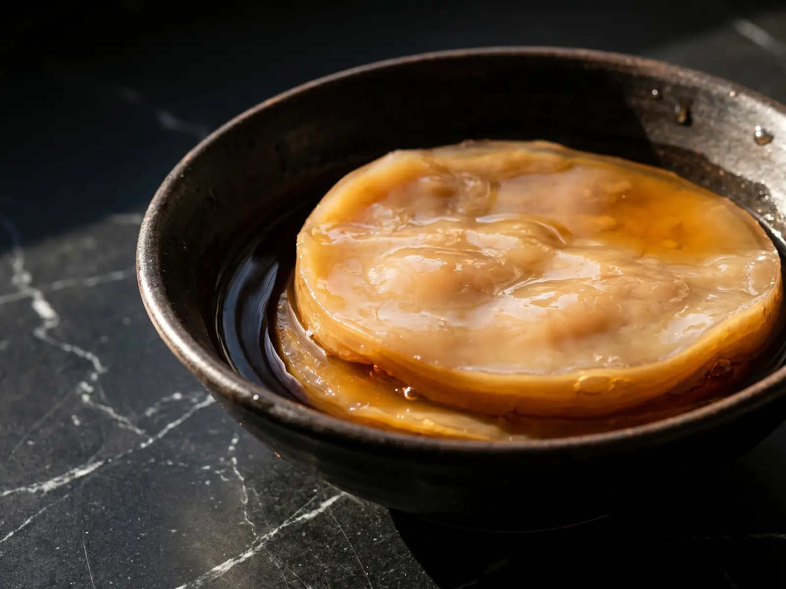 kombucha que es Cuenco con té oscuro y disco SCOBY beige sobre mármol negro con luz natural intensa.