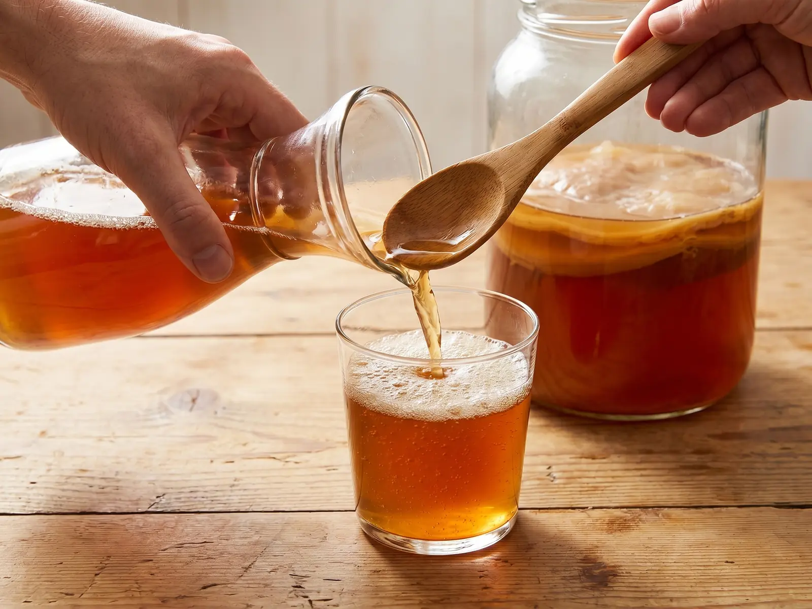 Mano sirviendo kombucha en un vaso de cristal con espuma fina sobre una mesa de pino.