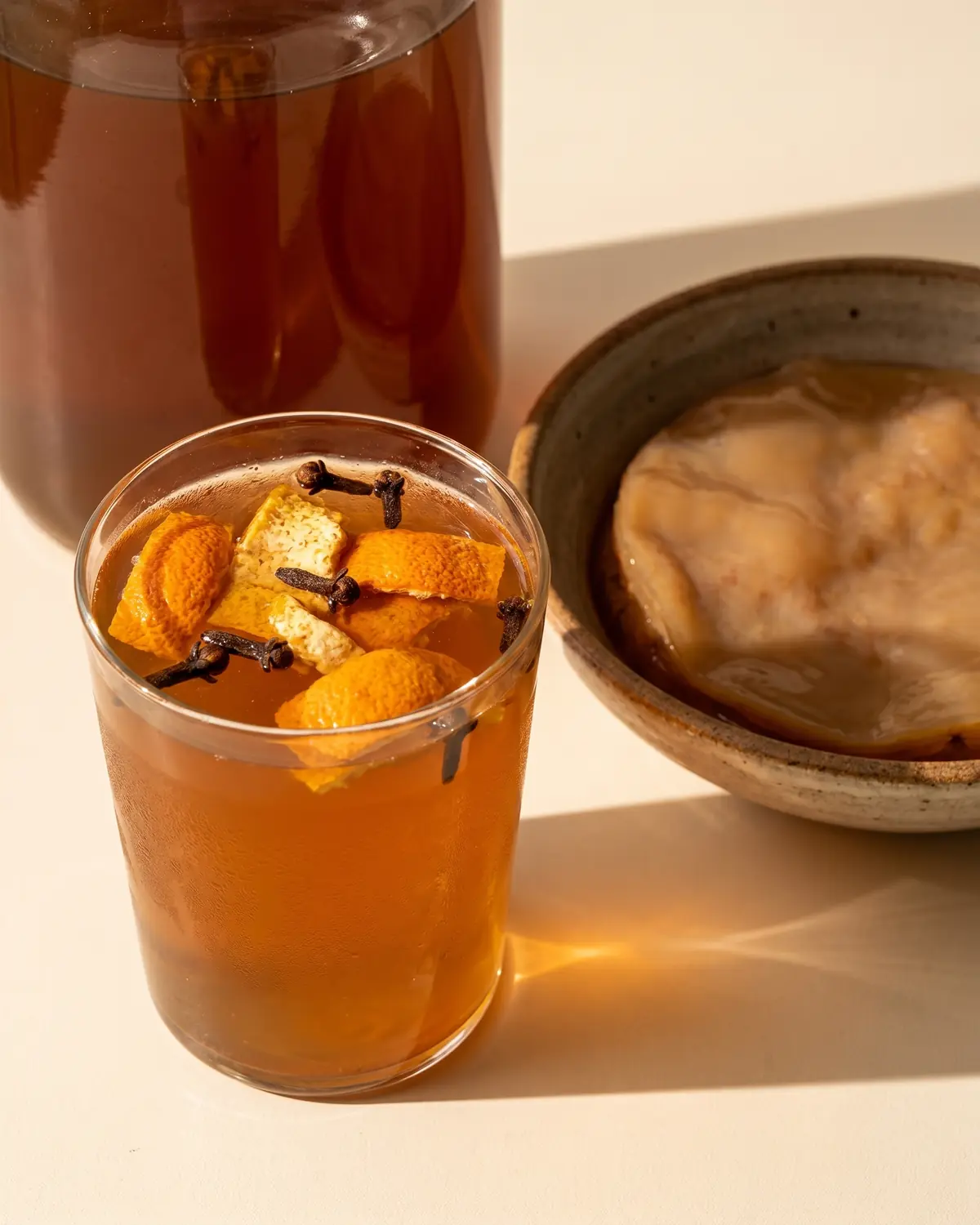 Vaso de kombucha &aacute;mbar con piel de naranja seca, clavo y una sombra suave de tarde.