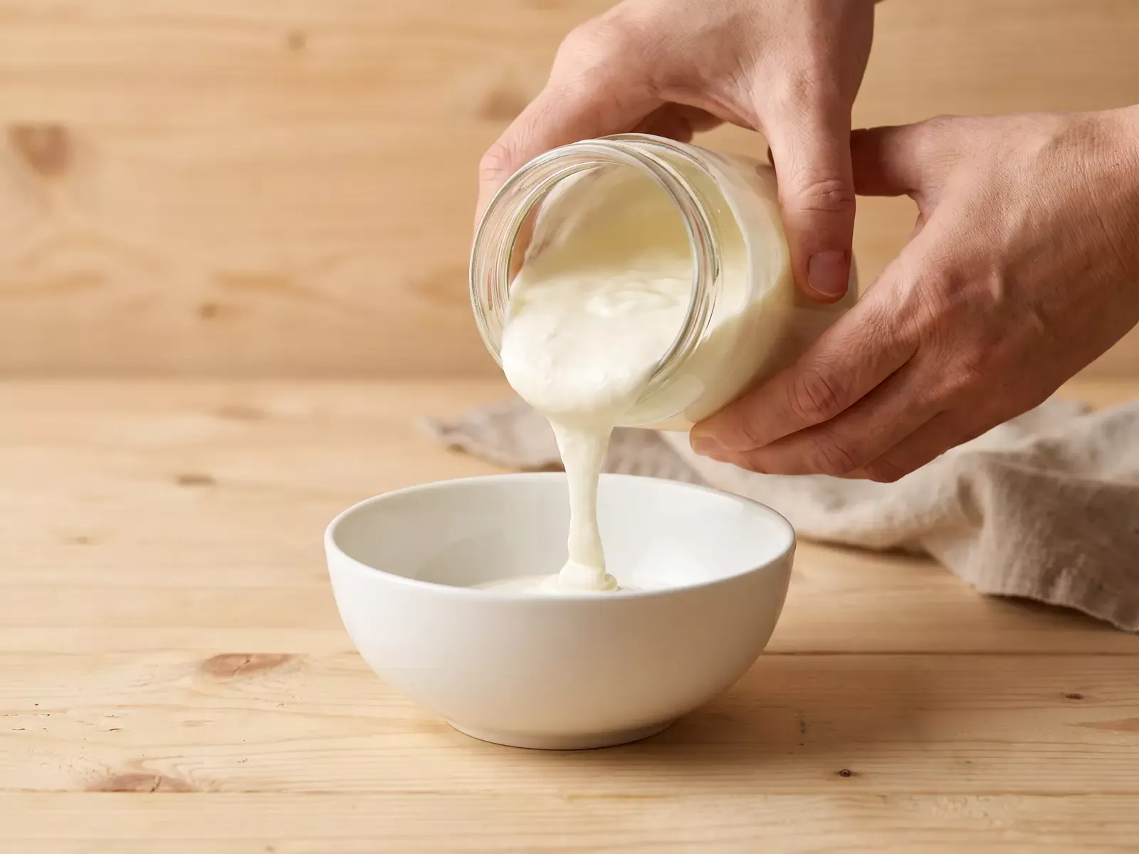diferencia entre skyr y kefir Dos manos vierten un yogur espeso desde un tarro hacia un cuenco blanco sobre una mesa de pino claro.